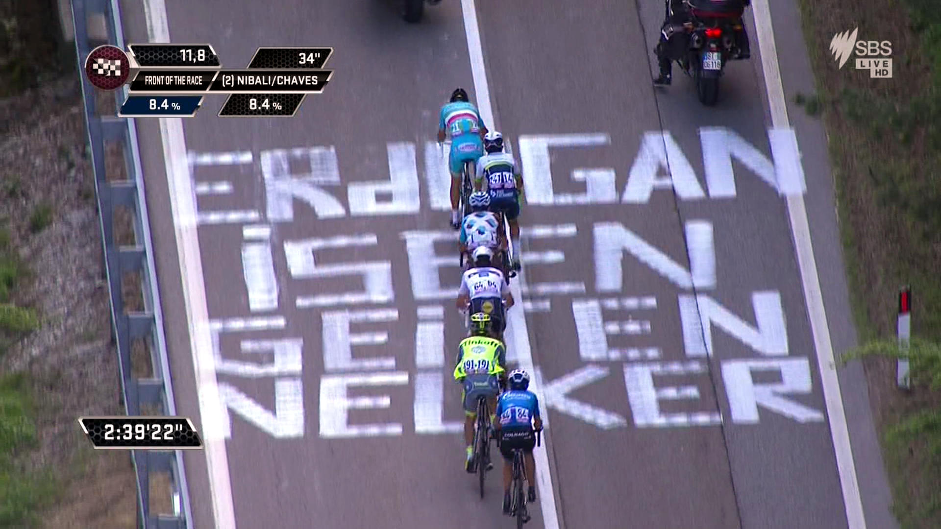 Riders in the Giro d'Italia pass over slogan on the road.
